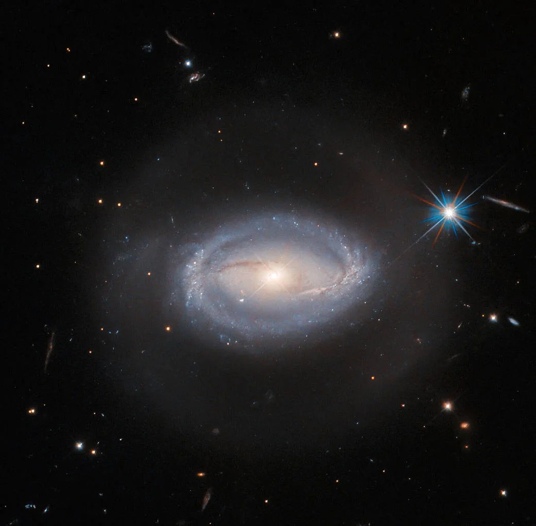 A spiral galaxy. It has two almost-straight arms coming from the left and right of the core that meet a starry ring around the galaxy’s edge. The ring is bluish in color, and the core is golden and shining. A faint halo of light also surrounds the galaxy. There is one bright star with many diffraction spikes, and a few small stars all around on a black background.