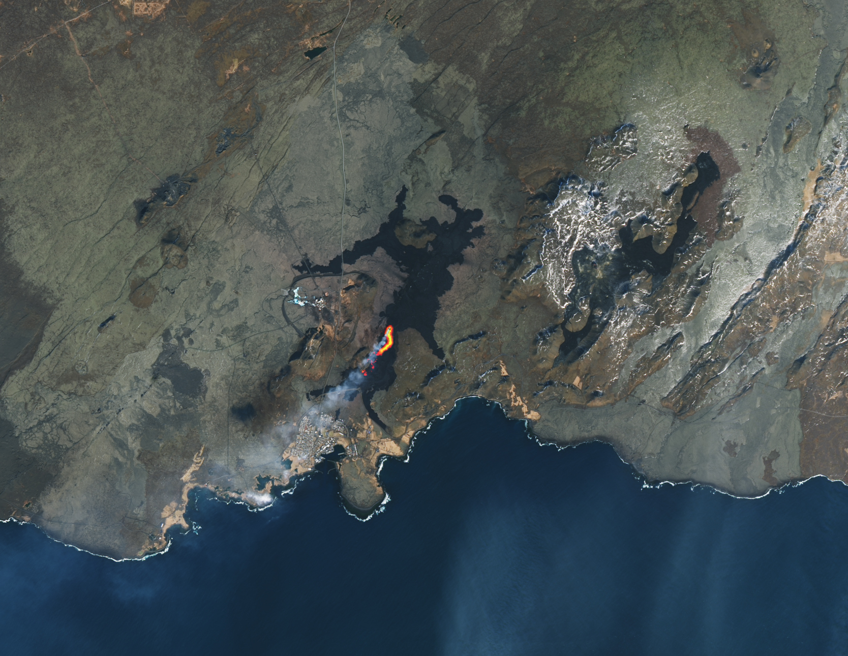 The natural color scene is overlaid with an infrared signal to help distinguish the lava’s heat signature. The active part of the fissure and the origin of a volcanic plume are apparent. This is seen in a bright orange slit centered in the image. Lava flows can be seen surrounding toward the top and right of the image where the land is largely empty. toward the bottom of the image is the coastline and the town of Grindavík along a small portion of the coast.