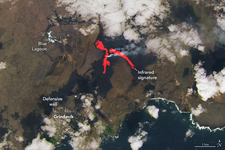 Satellite image of Reykjanes with infrared overlay to highlight the lavaflow shows two flow origins in a bright white with lava rivers coming from each. The land immediately surrounding this lava flow is dark black from previous lava flows. Clouds occupy the top center and right of the image and ocean the lower portion of the image with a population center on the coast.