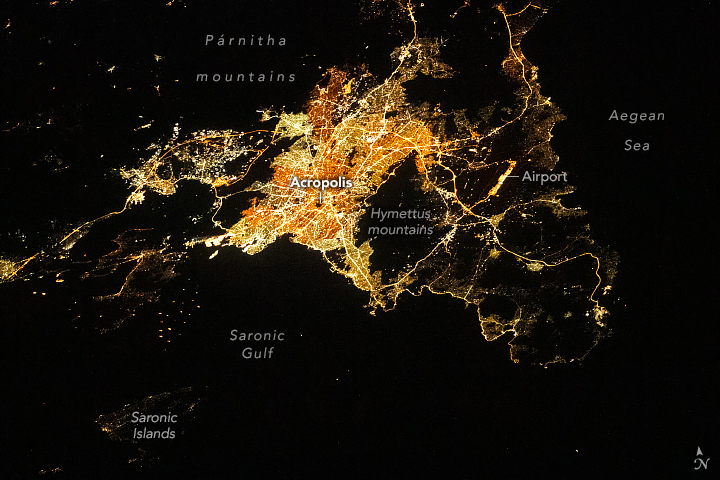 Aerial image of light illuminating Athens at night, a lone dark spot on the city indicates the Acropolis, but veins of light follow the streets out of the city and into other towns, illuminating the coastline faintly.