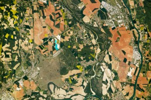 The Rhône River skirts the perimeter of the image and is joined by the Ain River near the bottom of the scene. The Rhône River appears dark blue, while the Ain River has a brown hue due to its higher concentration of suspended sediment. The river floodplains include meander scars and oxbow cutoffs, several of which are filled with dark green vegetation. Angular agricultural fields range from tan to green and vary in color due to primary crops, such as wheat, barley, rapeseed, corn, and sunflowers. Reddish agricultural plots indicate recent planting or tilling.