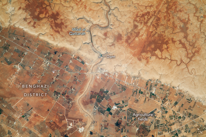 A dammed river splits this image down the middle, above the dam the land appears to be more challenging terrain, with dark valleys sprawling in all directions. In the lower portion of the image the land appears universally darker and hosts numerous agricultural fields.