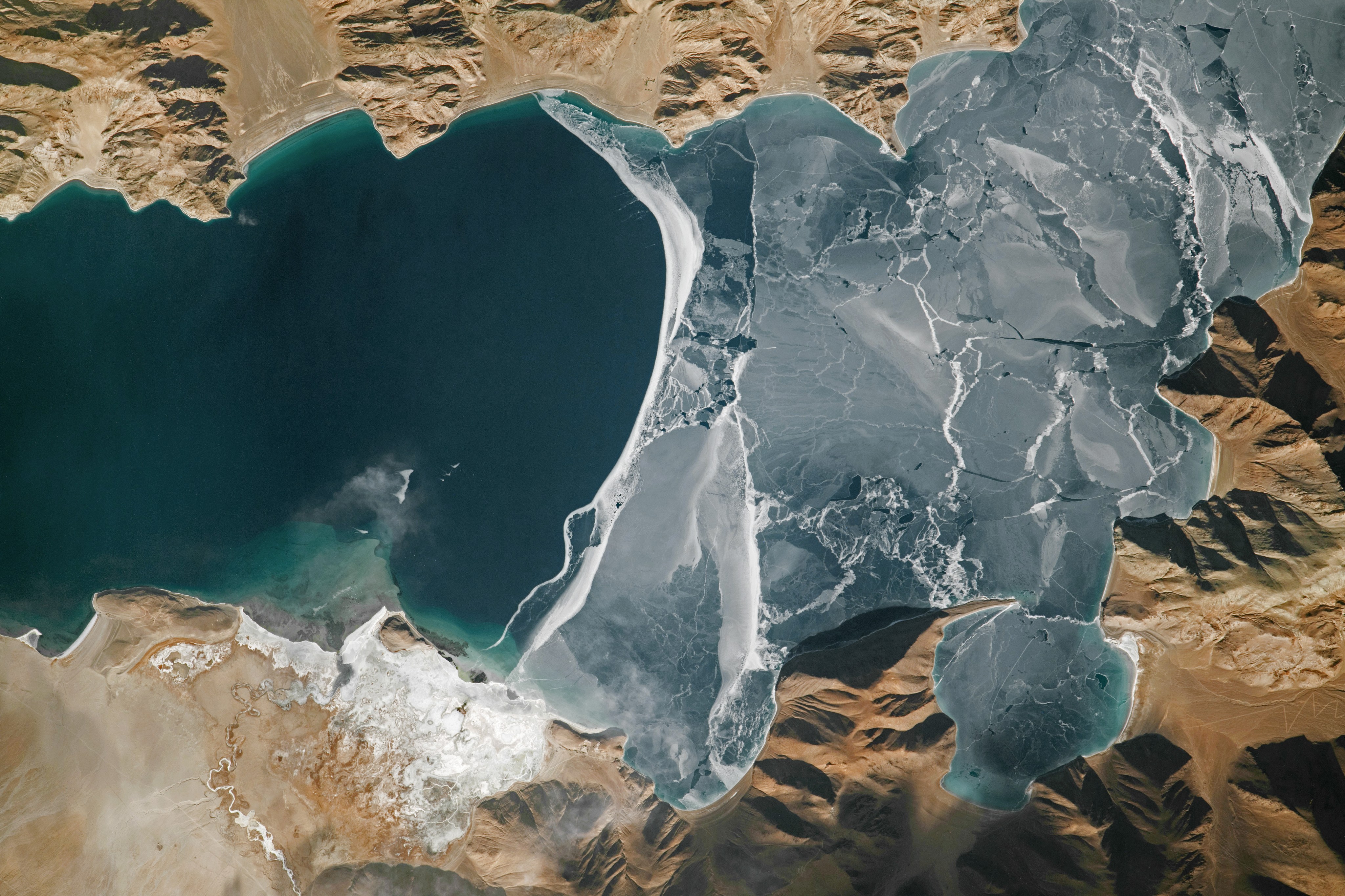 In this winter scene, a sheet of cracked ice tops part of the lake (right side of the image). On the left side, open water appears blue. Shallow water offshore of a delta, formed by a small, winding river, appears green. Several roads cut across the landscape, including one with switchbacks that ascends a steep slope.