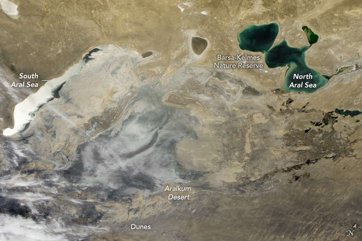 This image of the desert from space shows a few seemingly small bodies of water in a sea of gray and beige landscape. At the center left edge of the image is a longitudinal body of water labeled the South Aral Sea. It extends to the top middle left of the image and is quite narrow. Two small pools exist between this and the North Aral Sea in the upper right, which is blue green in color and is primarily composed of three connected rounded pools. At a right angle to both the land appears gray and aberrant from the surrounding desert sands.