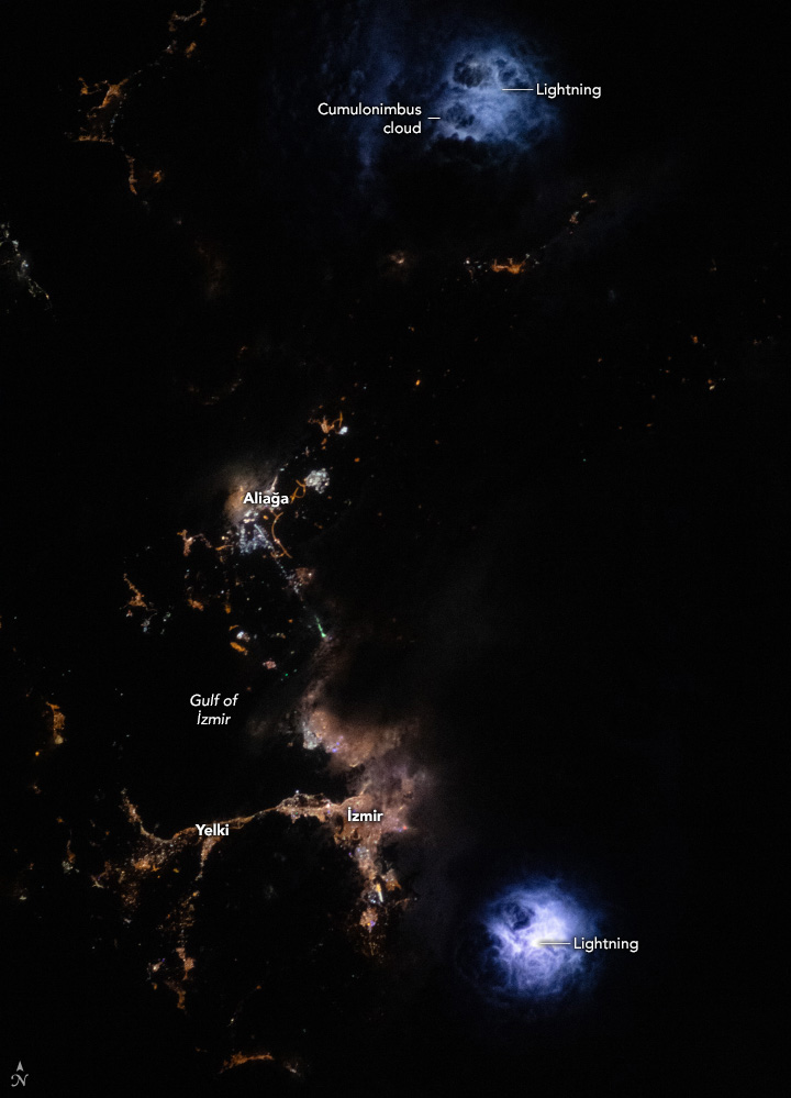 Webs of blue light appear in two places on the image, top right and bottom right, as lightning illuminates the night clouds. In the lower left of the image lights of cities are visible, but the dark cloud obscures the natural boundaries of those city lights edges.