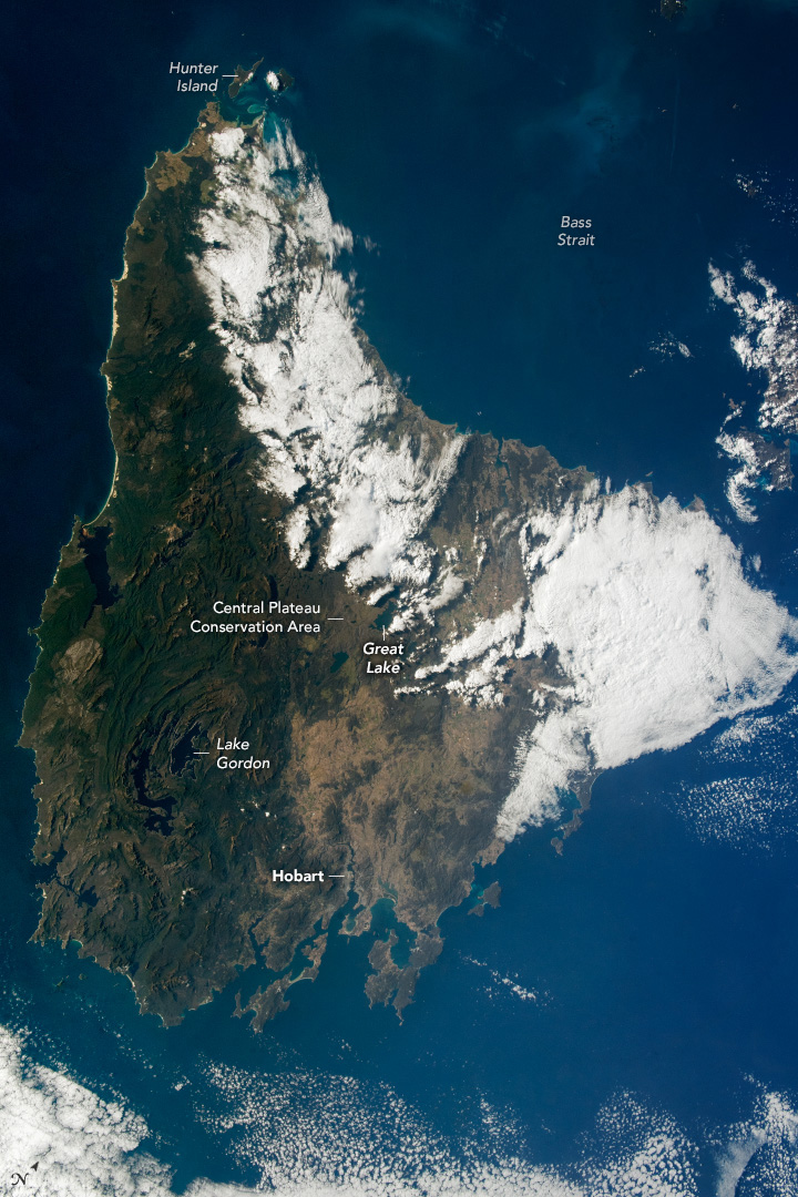 The island appears surrounded by a neutral shade of blue ocean. The island has two large regions spanning away from the center toward top and right of the image. The left of the island appears to be mostly green with the right side more brown in hue. The upper and right protrusions from the center of the island seem to be significantly cloud covered and appear white.