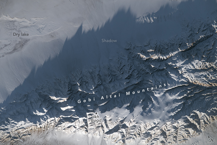 Mountain peaks span the entire bottom of the image, but also reached the upper right of the image in a steady diagonal. The lower portion of mountains appear bright white, illuminated by the sun, but cast a shadow on most of the rest of the mountains as well as the snow covered plain above.