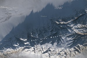 Mountain peaks span the entire bottom of the image, but also reached the upper right of the image in a steady diagonal. The lower portion of mountains appear bright white, illuminated by the sun, but cast a shadow on most of the rest of the mountains as well as the snow covered plain above.