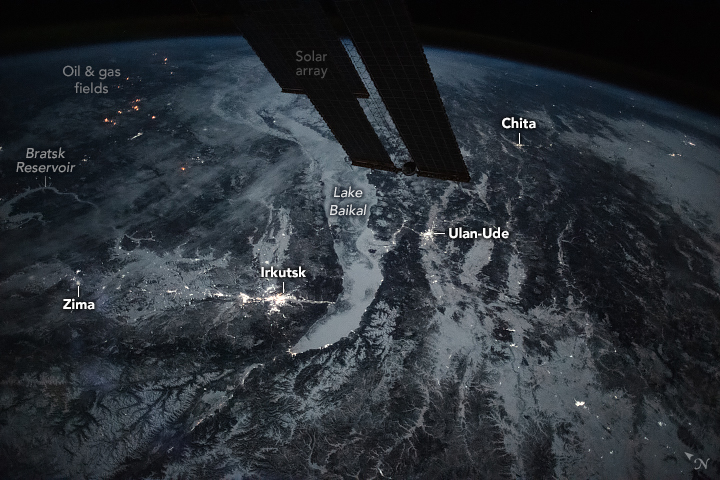 The image from the the International Space Station shows the solar arrays interrupting the top of the view of Earth. The landscape shows significant white reflective regions due to snow coverage. There are are also some city lights, most notably Irkutsk, dotting the landscape.