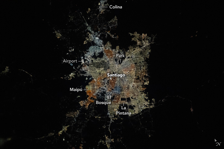 The dark image shows the night illumination of the city of Santiago. Colina lights are at the very top of the image, detached from the heart of Santiago. on the left of the city the Airport is visible as slightly separated, but brightly illuminated. On the upper part of the city a dark region is labelled as a park. Maipú is in the lower left, El Bosque the lower center and La Pintana is just right of El Bosque.