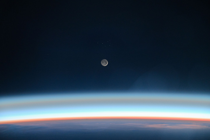 The glowing atmosphere of Earth appears at the bottom of the image. Above that, centered in the image, is the moon appearing mostly in dim light, with only a sliver appearing brightly illuminated.