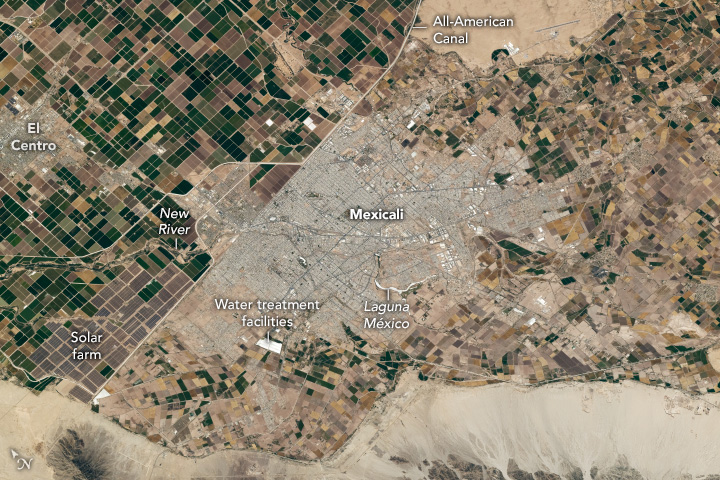 The beige city streets are surrounded by farm fields before giving way to sandy terrain. To the right the land plots are less regular compared to those on the left of the city, which are neatly gridded. The farm land to the left is also darker in color.