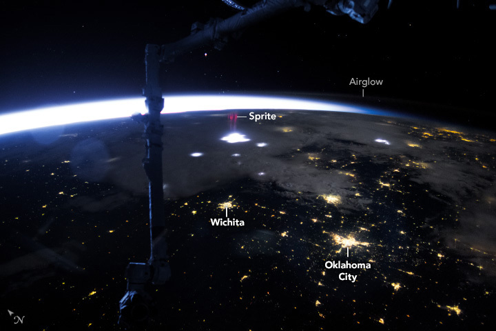 A view of the United States at night from the international space station shows equipment from the ISS in the foreground. On Earth a lightning storm brightly illuminates clouds in spots and city lights are visible in clear regions. Above a bright cluster of clouds a purple spot, labeled a sprite, is visible in higher atmosphere.