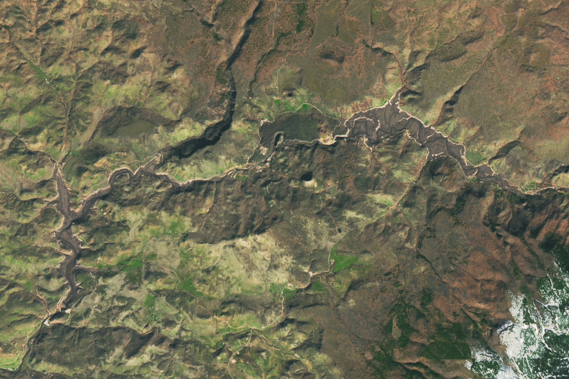A river enters the photo at the top right of the image, meandering through the accumulated sediment of what was previously Copco Lake, created by the Copco Dams. The river proceeds to cut its way through the craggy landscape that is covered in light green and brown vegetation. Numerous scars on the landscape indicate where rainwater collects and descends to the river The river enters another silty basin that was the Iron Gate Reservoir