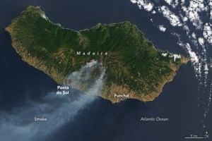 Satellite image of an island emitting smoke from the island's center which drifts toward the bottom left of the image. The island is primarily a deep green color with the coastline closer to the bottom showing much more brown coloration.