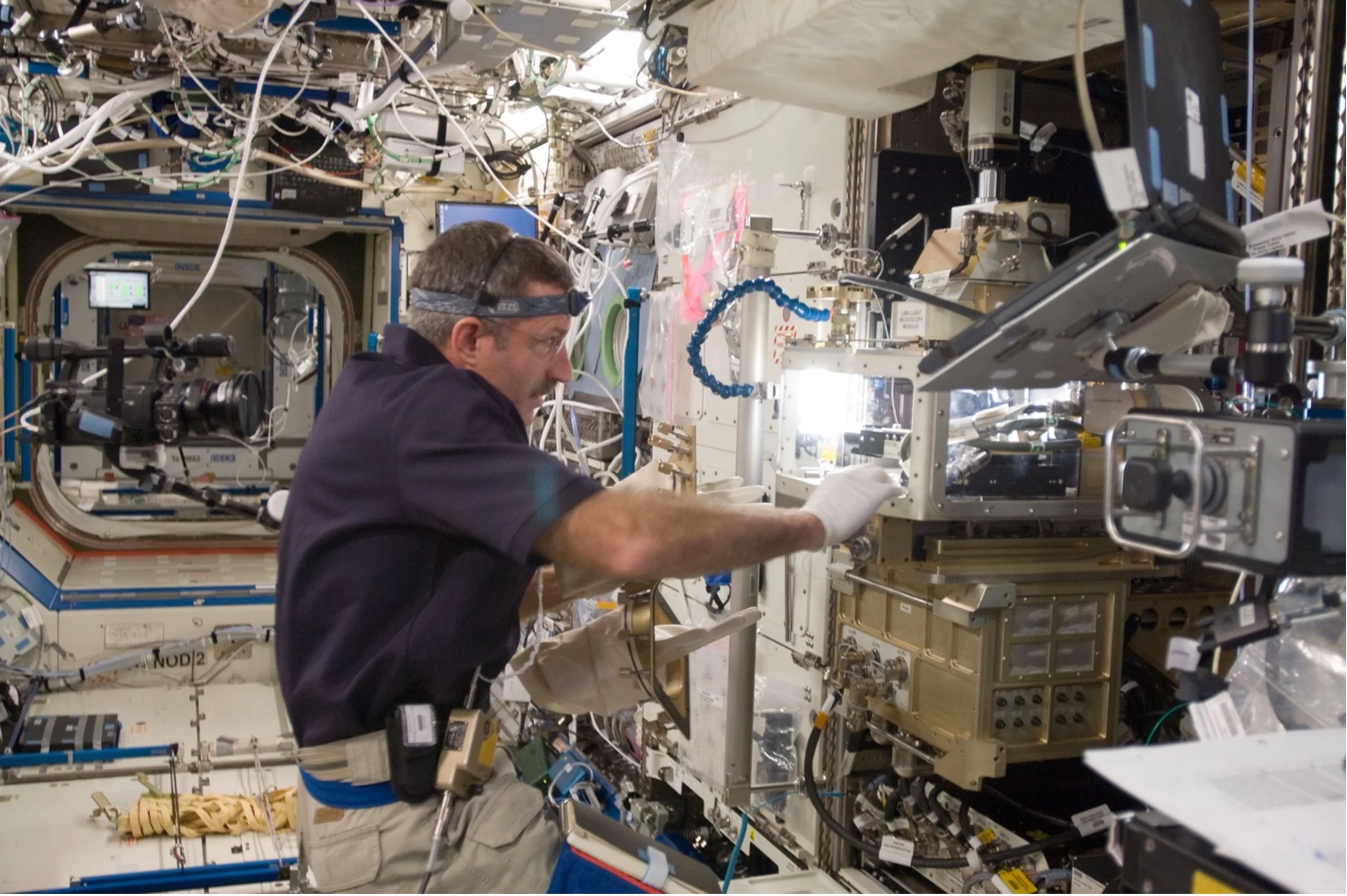 Astronaut inside the International Space Station holding onto white framed box with bright open area containing black wires and other mechanics inside.