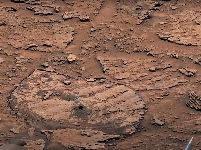A red-brown crusty and rocky surface is shown with a drill hole at the bottom center.