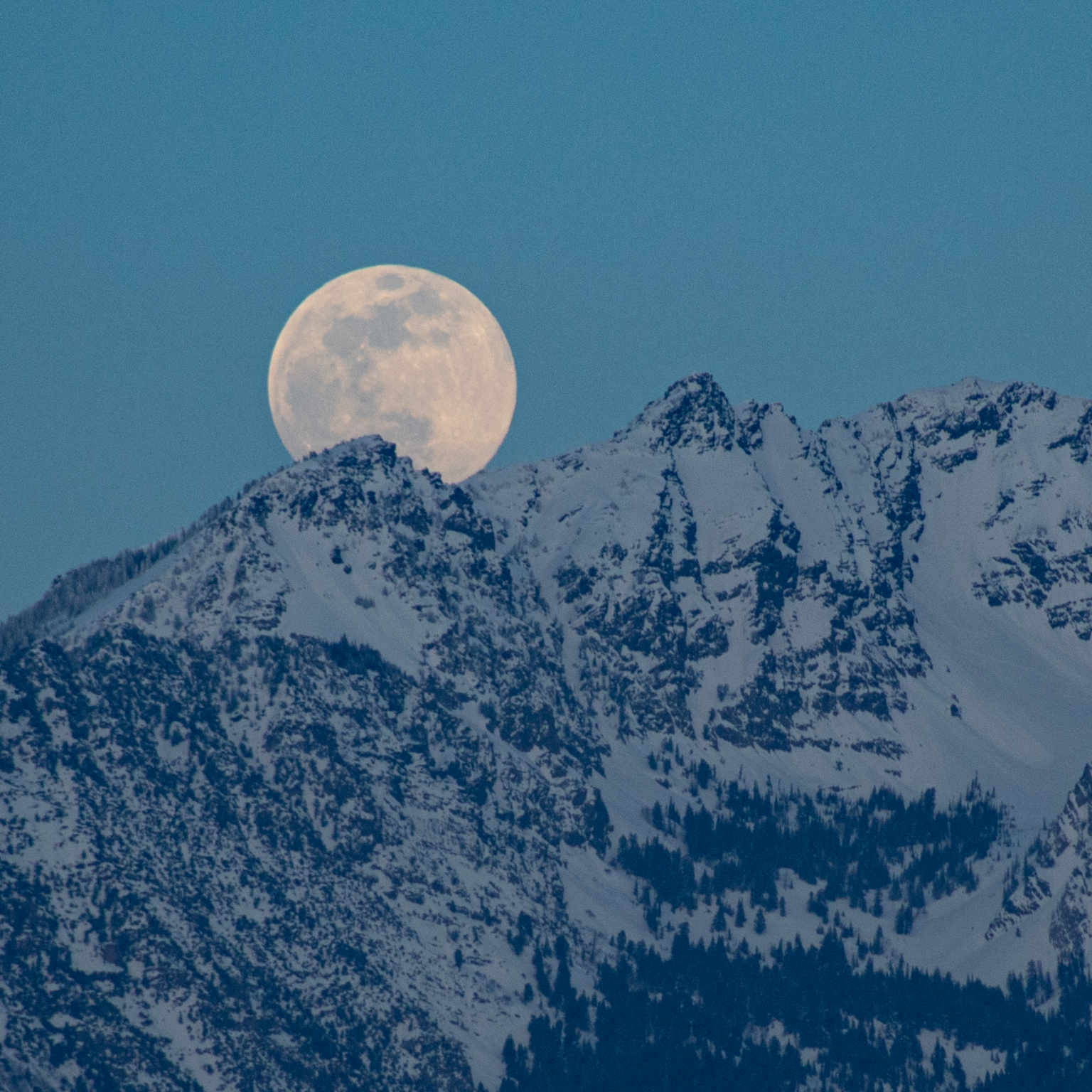 A full moon rises above snow-capped mountain peaks in this chilly image.