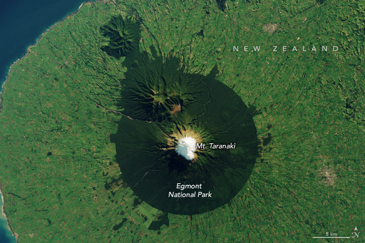 Mount Taranaki, capped white with snow, is the centerpoint for Egmont National Park. The borders of the park are clearly discernable from space as the land goes from primarily being the light green color of farmland to dark green forested in a perfect ring around the mountain peak. Smaller peaks to the upper left of the image are also forest covered, and some sections around the park remain forested as well, but the land is otherwise, primarily, light green.