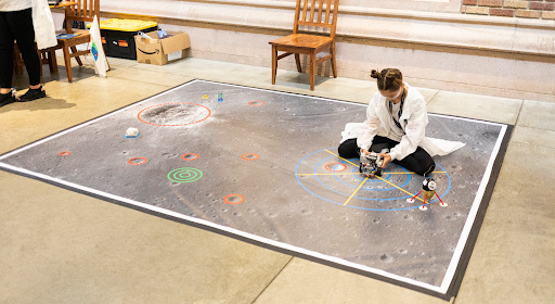 Photo of a young girl sitting on a large mat with an image of the surface of another planet that has various elements highlighted with red, green and blue circles.