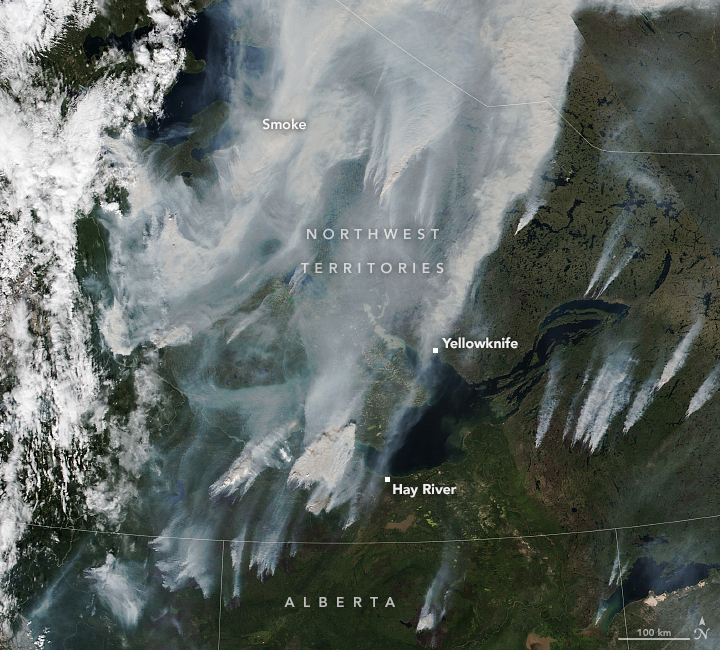 Satellite image of green landscape spotted with bodies of water. There is cloud coverage on the left the center is covered in smoke, and there are streams of smoke from smaller fires on the right.