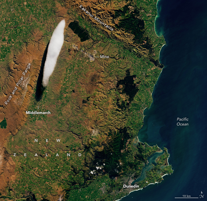 Coastal New Zealand is shown, the land is depicted left and the Pacific Ocean right. At the left edge of the image is a rusty brown colored land labeled Rock and Pillar Range. It ends before reaching the top of the image where the Kakanui Mountains descend from the top center diagonally down to the coast. A large cloud sliver appears between these two high elevation formations, casting a visible shadow onto the land below. At the bottom of the shadow the town of Middlemarch is labeled. Right of the cloud and below the Kakanui Mountains a mine is labelled and the color of the land is different in that spot. on the Southern coast the City of Dunedin is labelled.