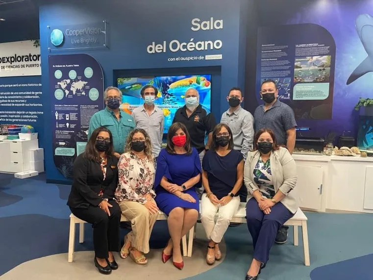 Photo of a group of 10 people wearing facial masks and sitting in front of an ocean display