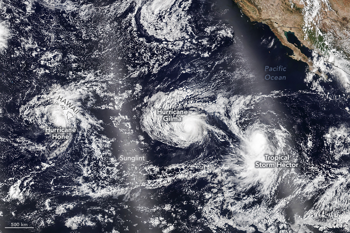 Dark blue ocean waters of the pacific dominate the image with significant cloud coverage scattered across the image. Centering in the image is a swirl of clouds labelled Hurricane Gilma, to the left, covering Hawaii is a mass of clouds labelled Hurrican Hone. Tropical Storm Hector is in the lower right which is less organized. The Southwestern United States and Mexico are seen in the upper right.