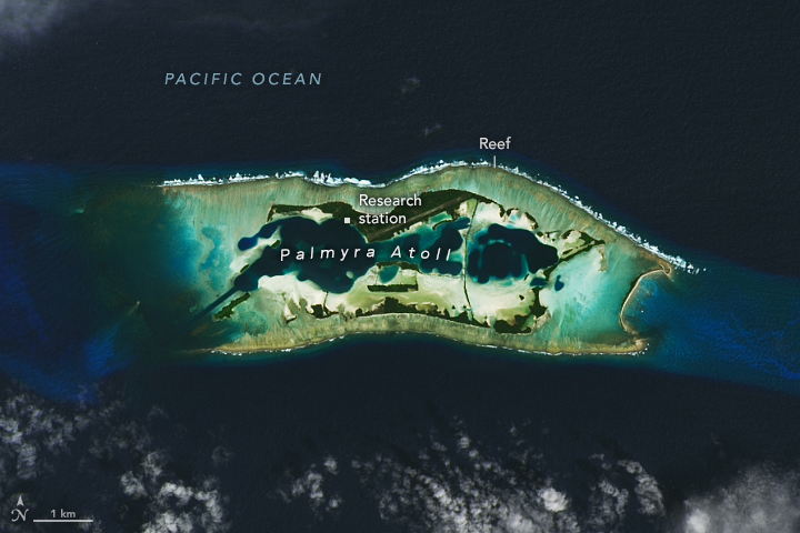 A strip of land and shallow waters centers in the image, aligned horizontal, surrounded by the dark blue of the Pacific Ocean. The atoll's interior boundaries also shows some dark blue waters.