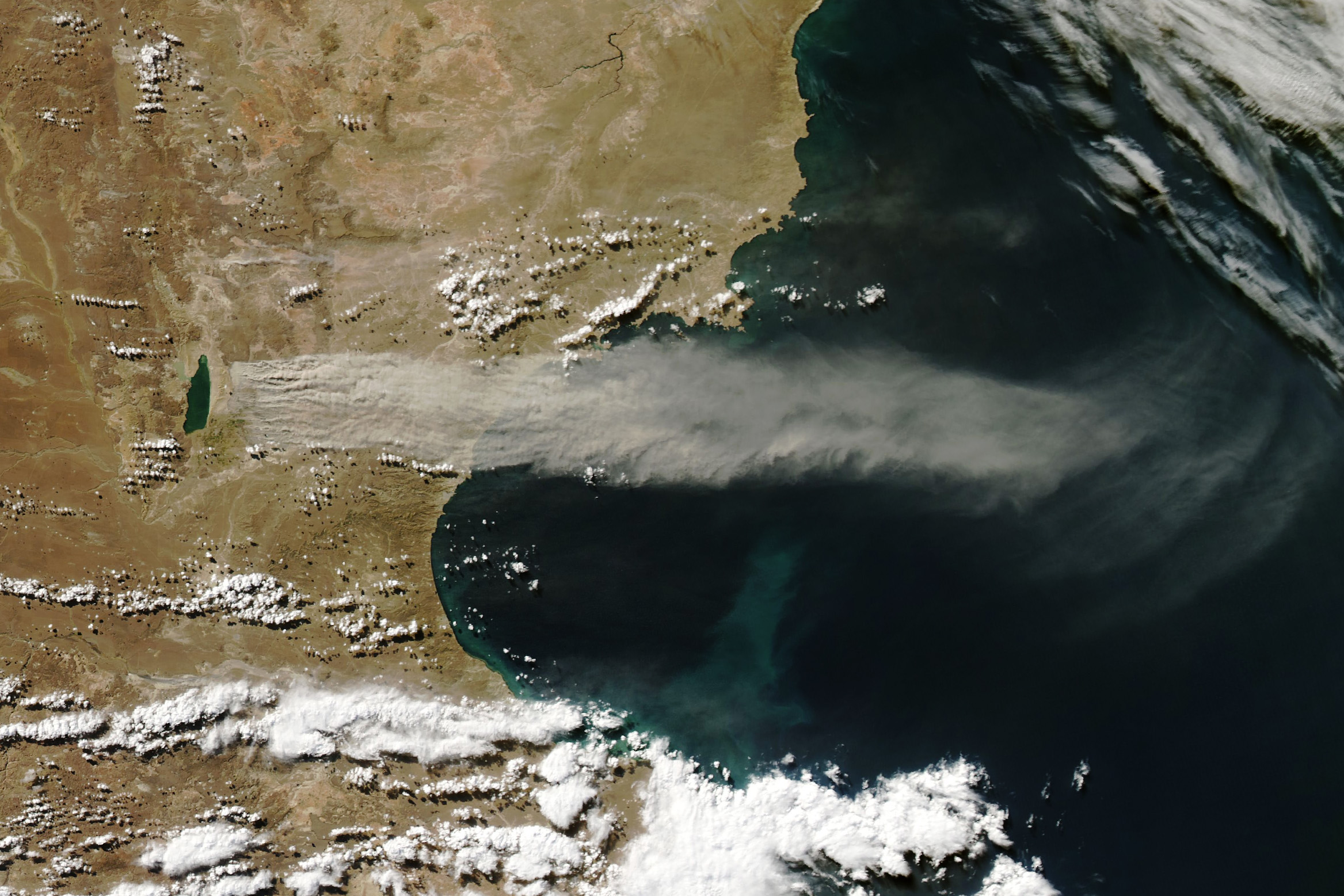 landmass of Agentina, left, and the Atlantic Ocean, right are featured around the San Jorge Gulf alcove in the coastline. Centered in the image is a horizontal stream of dust that stretches from the inland Lago Musters fully across the gulf and dissipates out in open ocean air. White Cumulus clouds are scattered south and mostly west of the San Jorge Gulf