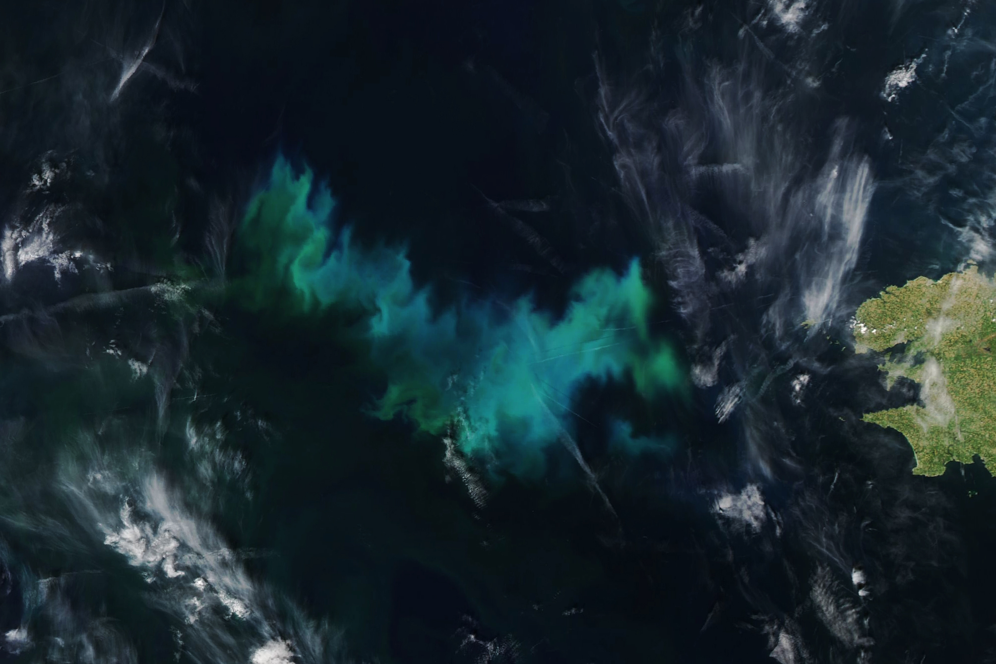Centered in the image is a disorganized patch of light blue and green color in the dark surrounding ocean. Some slight swirling within the colorful area is visible, thin windswept clouds are scattered primarily in the lower left and the mid and upper right. on the right side of the image is a bit of landmass, France, that seems to be largely green, but polka dotted with yellows throughout.