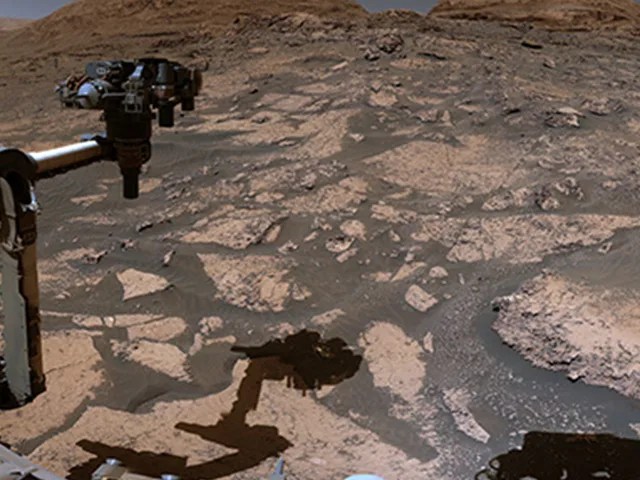 A robotic arm is shown protruding at the right, over a dark brown and light brown sandy and rocky surface.