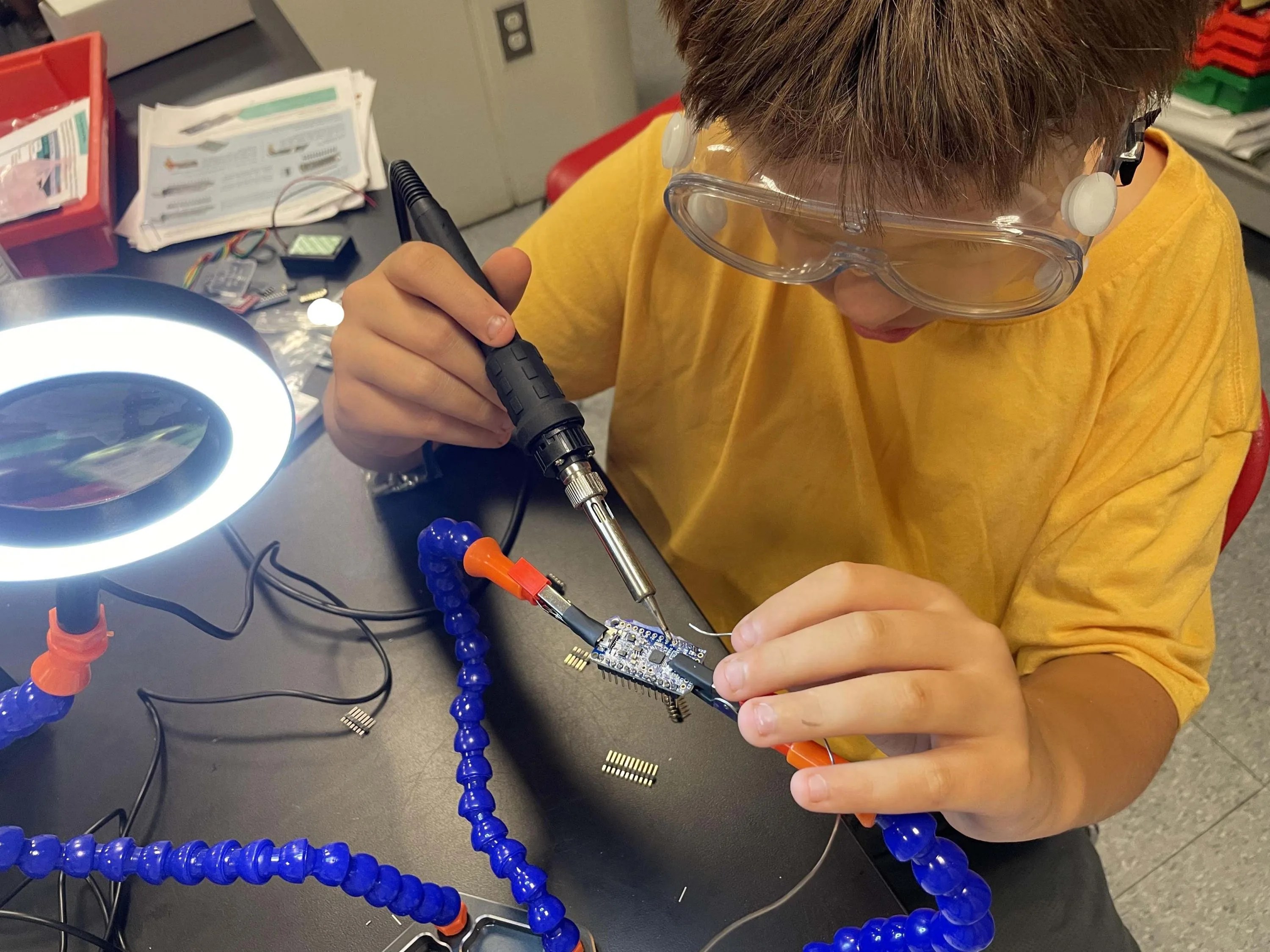 Photo of middle school camp attendee soldering the microprocessor used in the rocket payload. The microprocessor is held by clips attached to moveable arms for ease of access.
