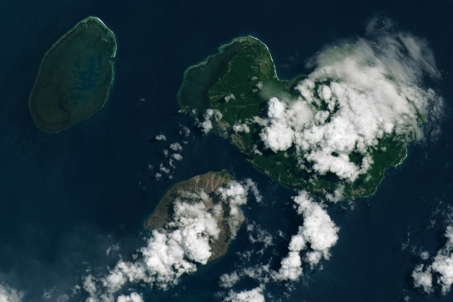 Ruang Island centers, it is brown in color above to the both right and left are other islands that are a vibrant green color. large white cumulus clouds span the image top right down past ruang to the bottom left.