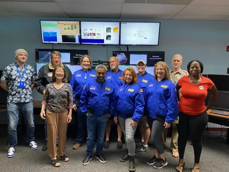 11 people, 6 wearing blue NASA jackets, in room with multiple large display monitors