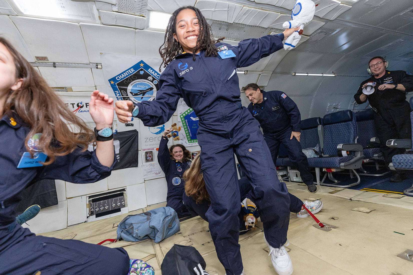 A group of individuals experiencing weightlessness inside a microgravity simulation aircraft. The participants, wearing dark blue flight suits with "Zero-G" logos, are floating and smiling, with one person in the center enthusiastically reaching out while floating mid-air. Others are interacting with floating objects or holding onto the sides of the aircraft, which is equipped with seats and various equipment in the background. The mood is lively, and the setting suggests a space-related educational or research experience.