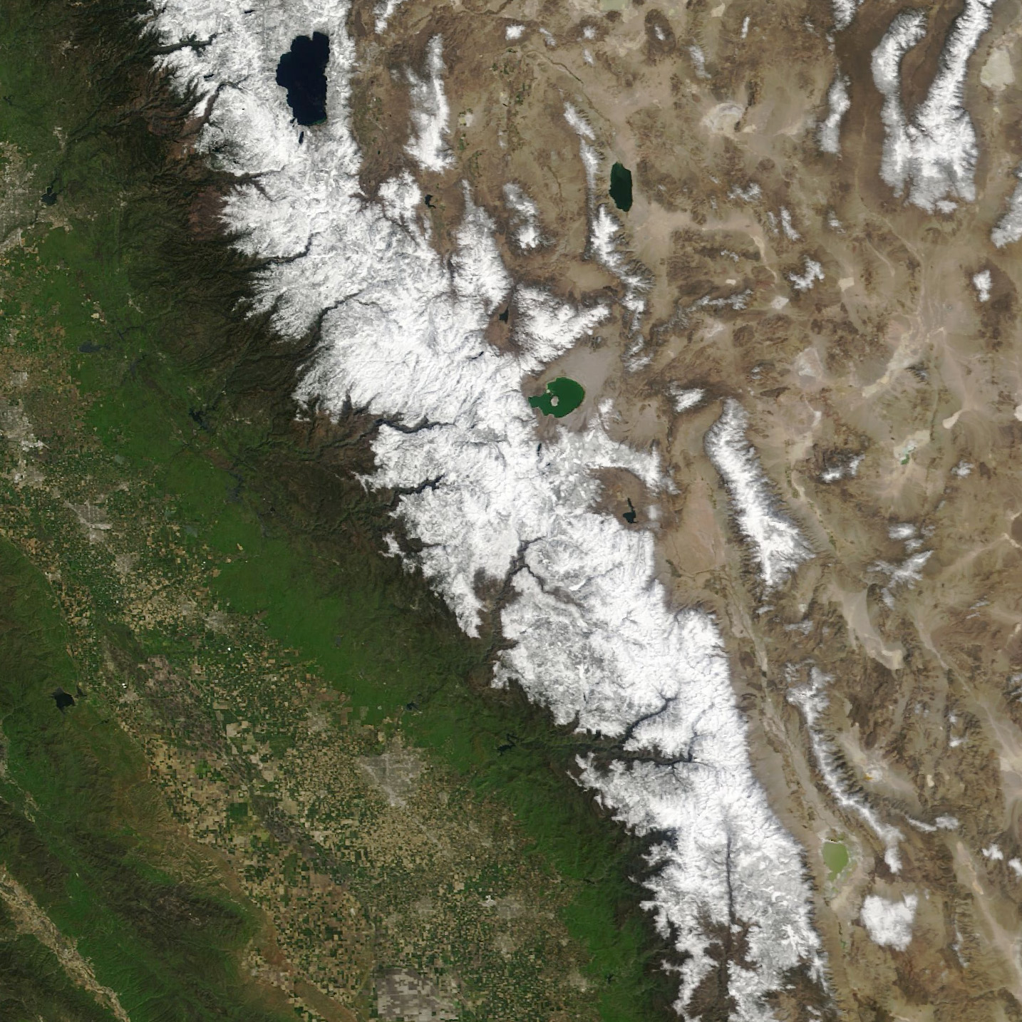 Satellite image shows the varied landscapes of central and eastern California. On the left is the verdant green San Joaquin Valley, cutting diagonally across the image from top left to bottom right are the Sierra Nevada Mountains which are significantly whitened by this winter's snowfall. To the right is the more arid and sandy climate, typical to Nevada. There are sporadic snow covered peaks.