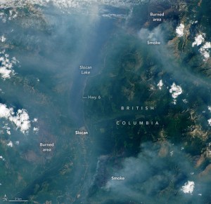 The image shows the valley—bounded by the Valhalla Range to the west and the Slocan Range to the east—filled with smoke. Slocan Lake appears near center, stretching vertically, as a dark poorly defined region through the haze.