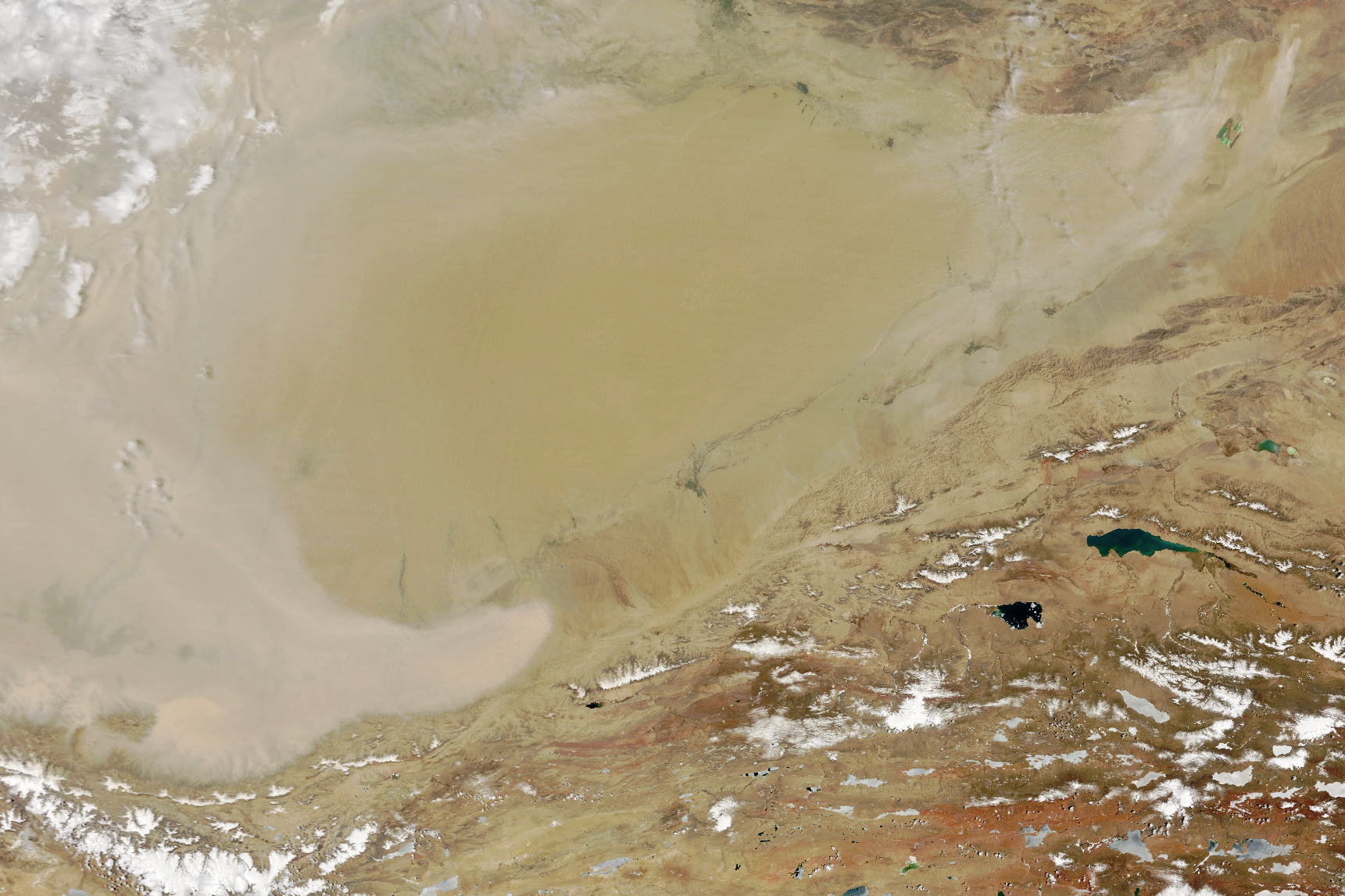 Satellite image of the light tan, nearly off-white, Tarim basin. There are some small lakes in the mountainous region seen lower right and those mountains span center right to lower left, speckled with snow and ice. In the top left there appears to be either ice or wispy cloud cover.