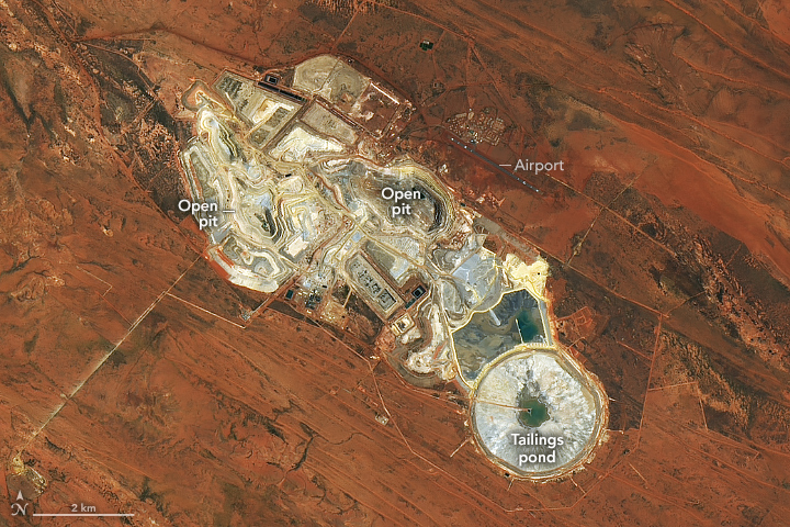 The rust red landscape is interrupted by an open pit mine that leaves the land gray and white. It stretches vaguely diagonally across the image, with a nearly perfectly round area toward the bottom right for a tailings pond.