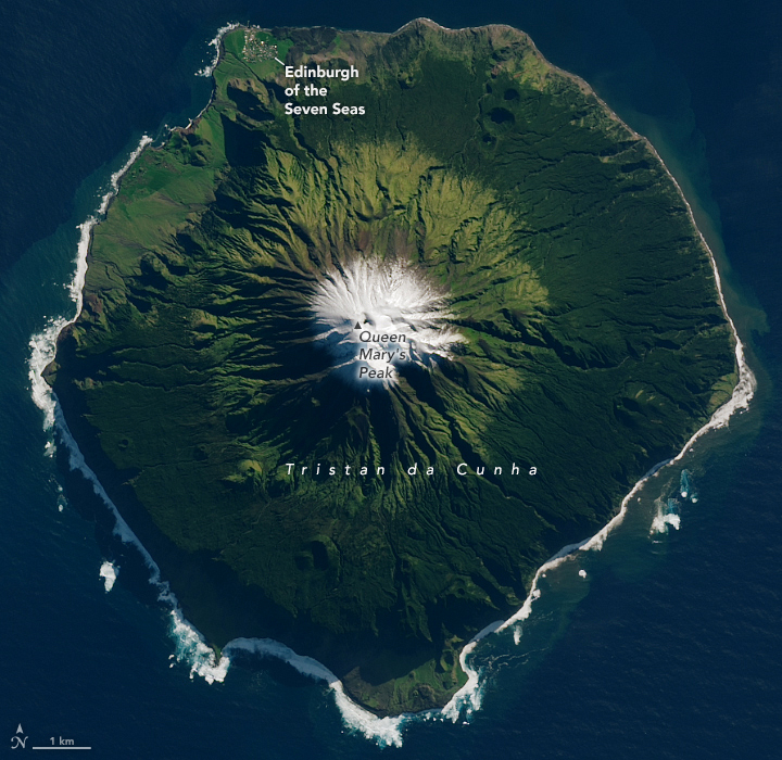 The image centers on a snow covered peak of a single fairly uniform circle of an island. The vast majority of the island is covered in foliage, the upper ring elevation just below the snowline is a lighter shade of green than that closer to the coast which appears a much deeper darker shade. The island shows a plateau along the coast in the upper left where the human population seems to have settled.