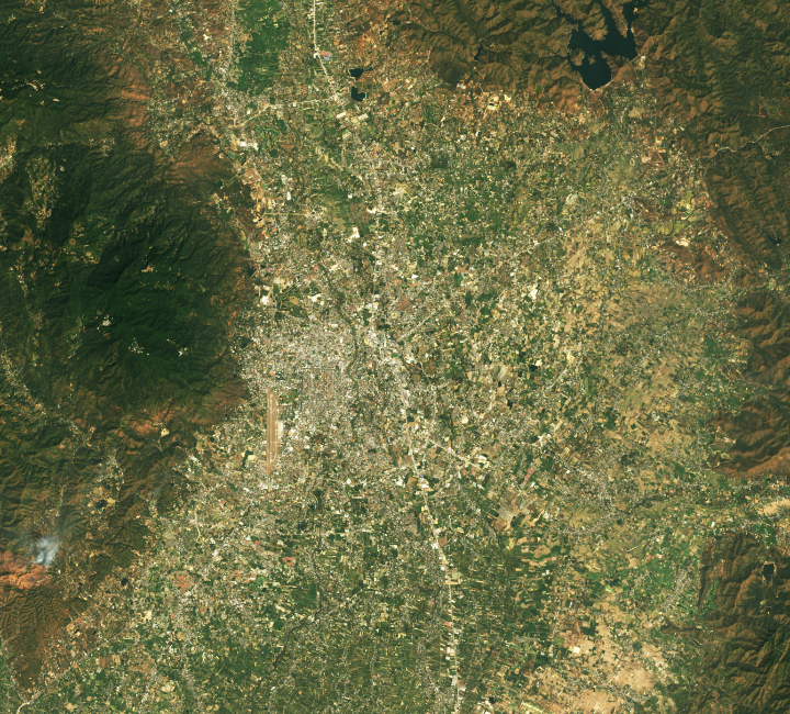 The image shows the sprawling city of Chiang Mai during day. The airport is visible to the lower left of the city center. further left is mountain Doi Suthep, lush green but in all other directions from the city center are signs of human habitations radiating outward broken up by what is presumably farm land.