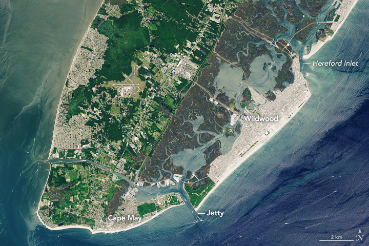 A peninsula extending from the top of the image shows light blue waters surrounding it. The left of the peninsula shows significant human habitation, but also significant green space, along the right of the peninsula are narrow inland waterways and a heavily occupied barrier island with a wide sandy beach.