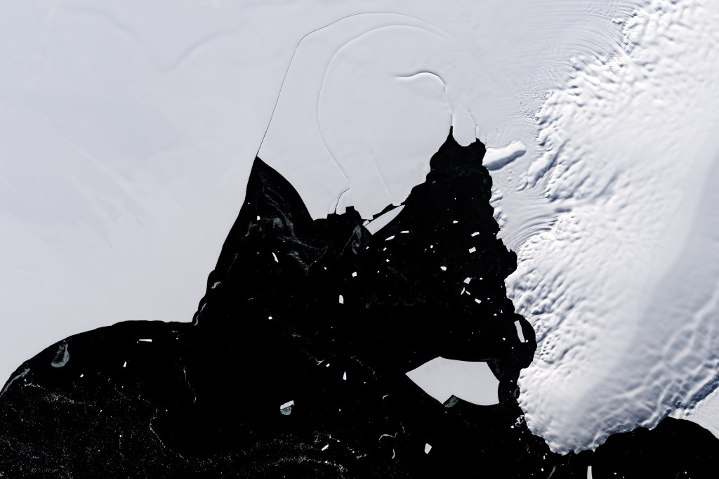 There image is primarily white sea ice with dark ocean at the bottom of the image along with sporadic icebergs. The- feature is the small hole in the Wilkins Ice Shelf that exposes the underlying ocean. This oddity has persisted for decades and is thought to be a rare phenomenon.