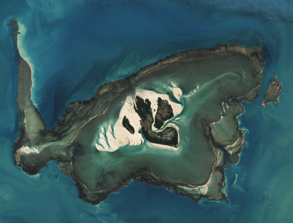 An island centers in the image, with clearly defined sandy shores in the bright blue Indian Ocean. However, there is a dark ring created by a reef around the island, some bright sand peaks above the water. There is also a sandbar going up the left side of the image.