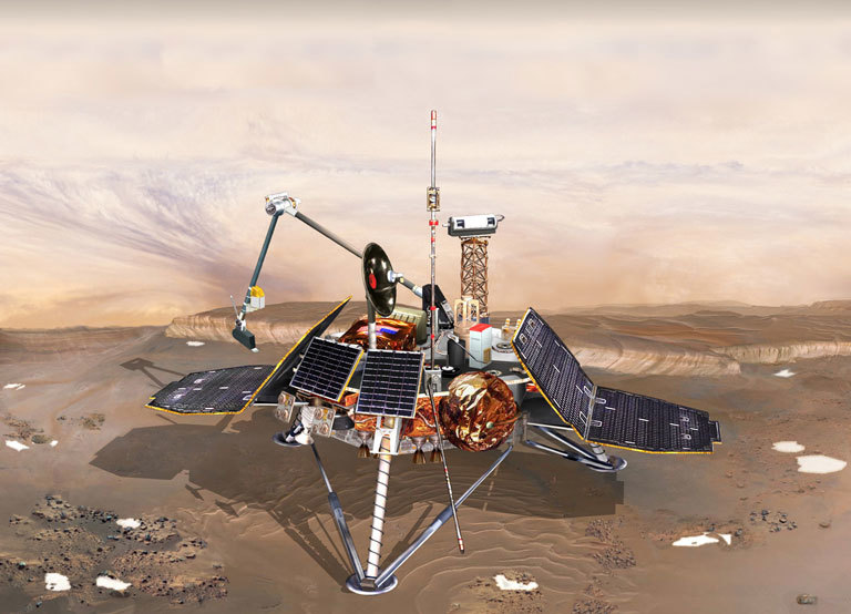 An illustration of a spacecraft lander on Mars, showing a thre-legged craft sitting on a desert landscape of brown and light orange soil dotted with patches of white ice. The craft .carries V-shaped solar panels attached to its laft and right sides, a camera atop a scaffold rising from the craft's top deck, and a spindly robot arm in back with a small scoop at its end.