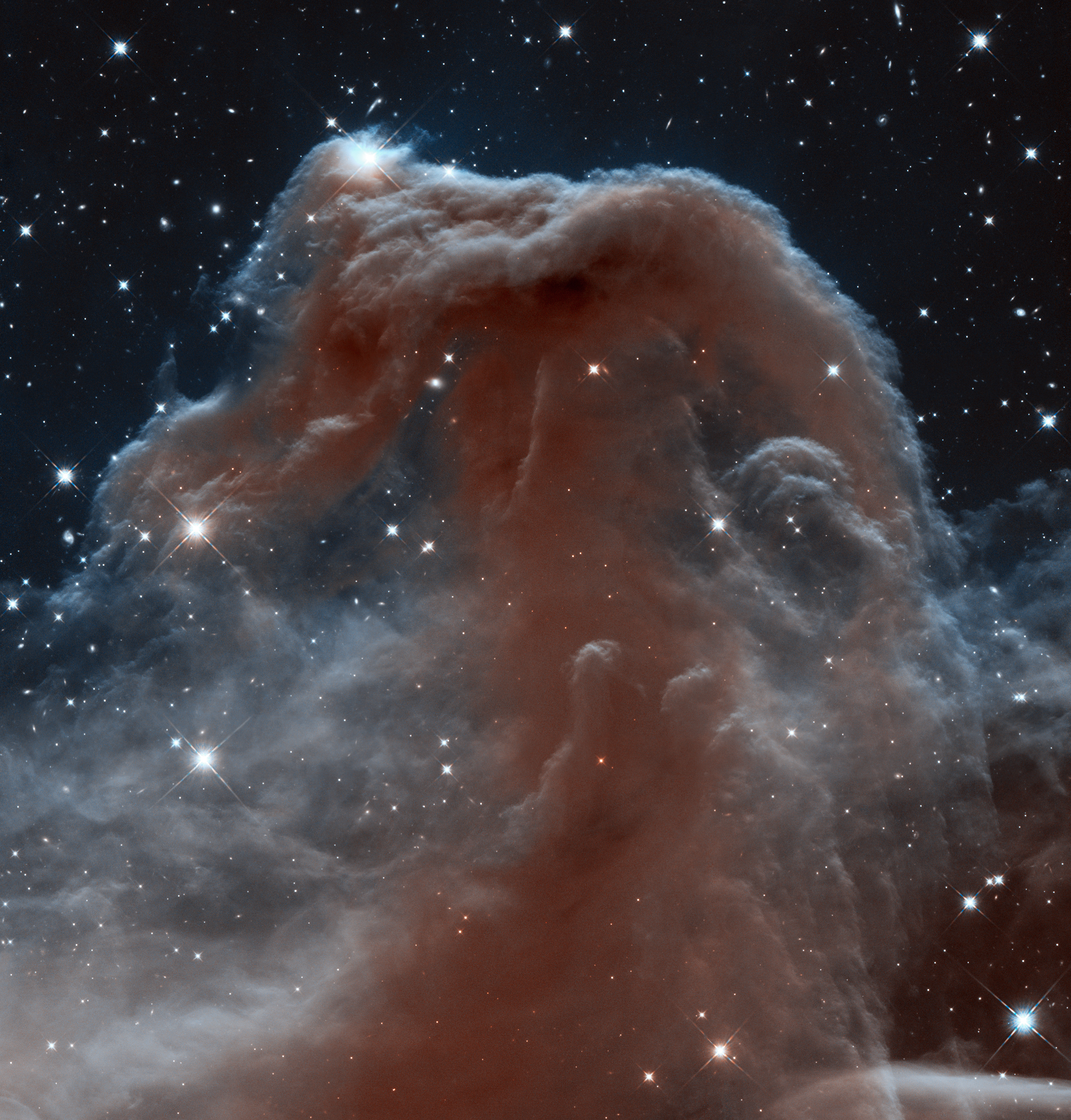 Large, dark red cloud of dust and gas surrounded by grey wisps of smoke and gas. Many stars in the black background of space.