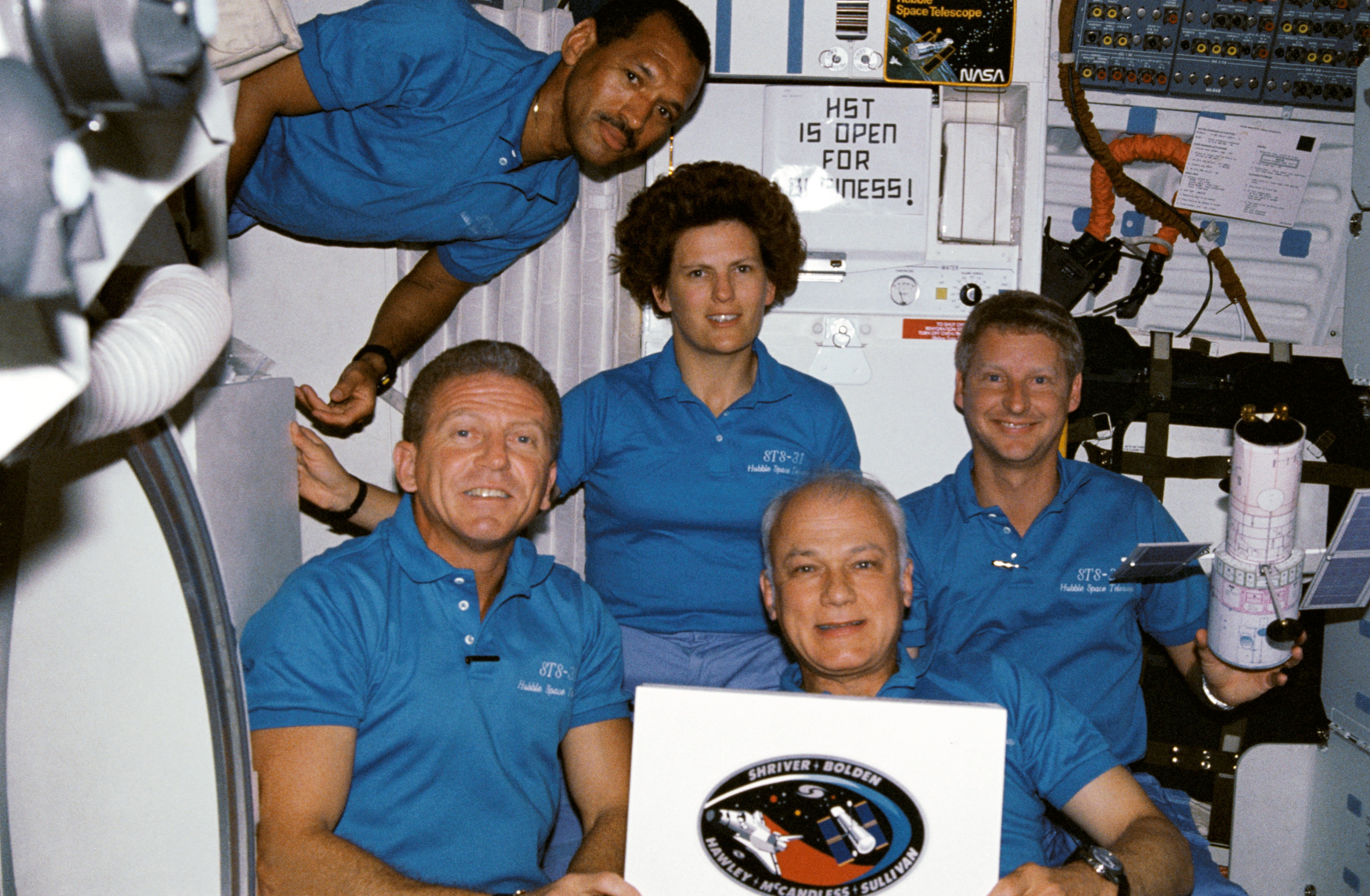 The STS-31 crew poses for a photo in the crew cabin in front of a sign that says "HST IS OPEN FOR BUSINESS!"