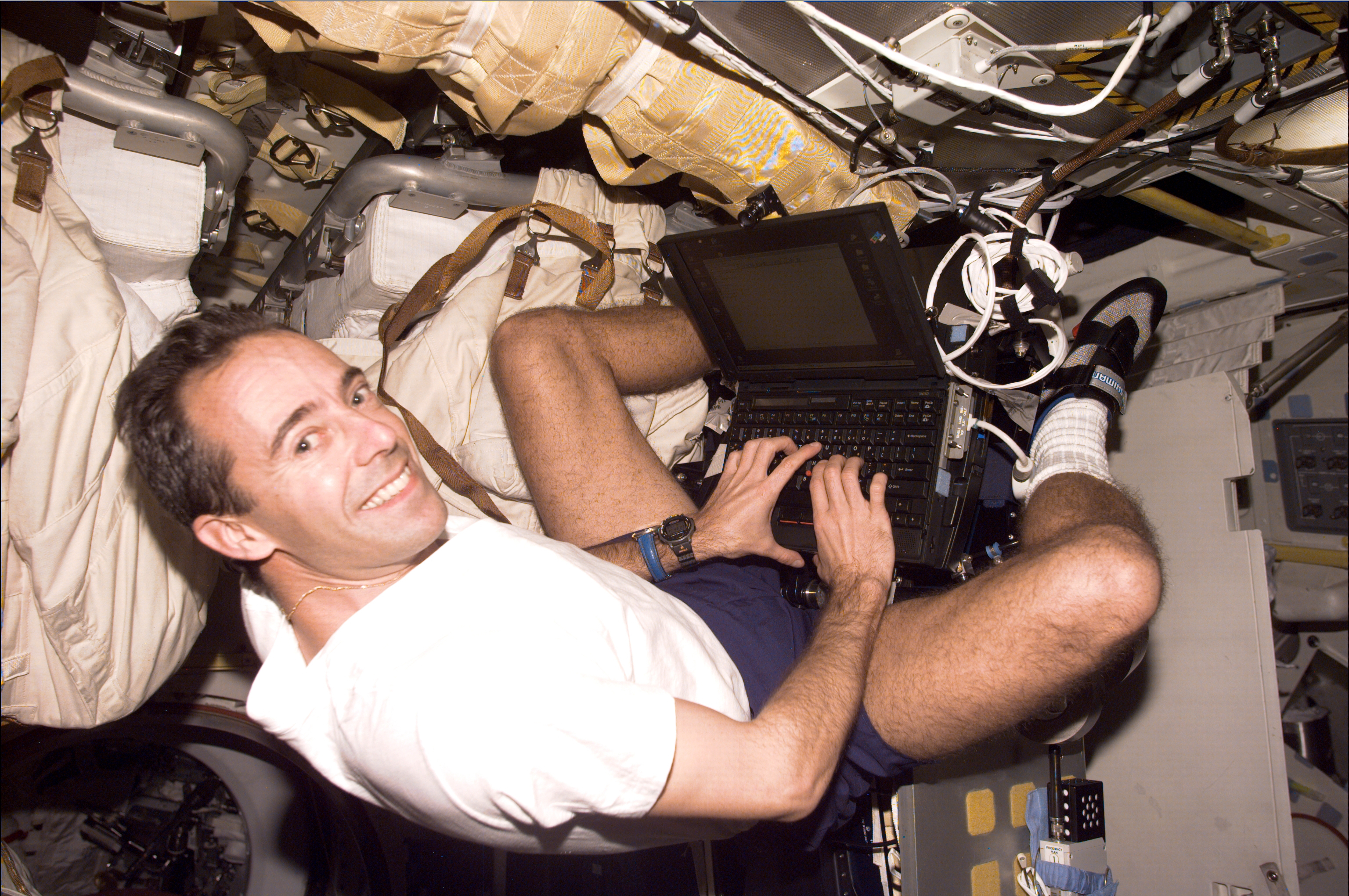 An astronaut in short sleeves and shorts works on a laptop in the crew cabin.