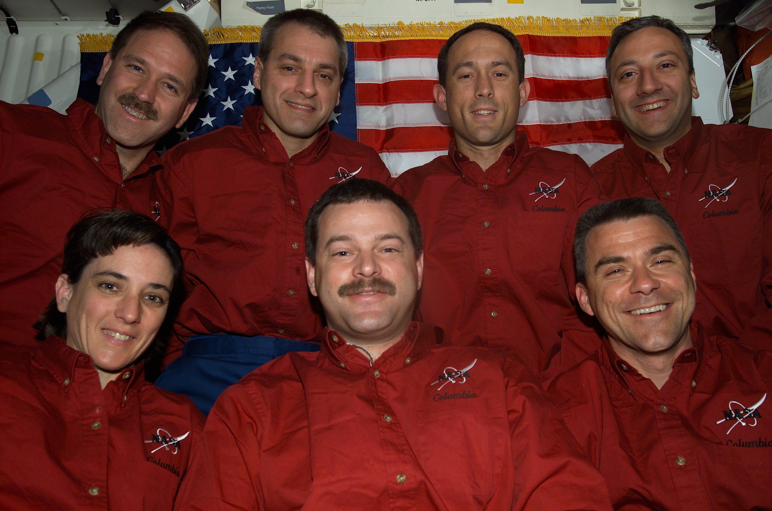The STS-109 crew poses for a photo in front of an American flag in the crew cabin of the shuttle.
